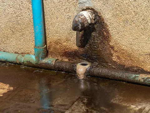 An old rusted water pipe leaking at a joint along the foot of a wall, with a rust stain spreading up the wall and water pooling on the ground