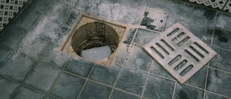 An open floor drain in a grey tiled floor with its metal grate lifted off and lying beside it, the tiles stained and scaled
