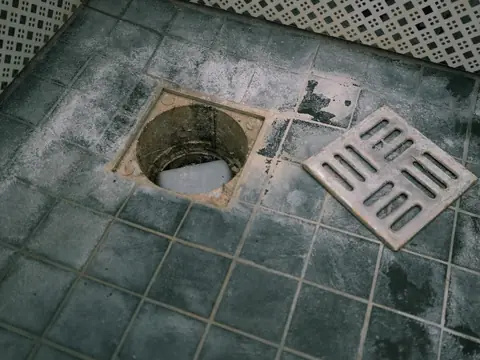An open floor drain in a grey tiled floor with its metal grate lifted off and lying beside it, the tiles stained and scaled